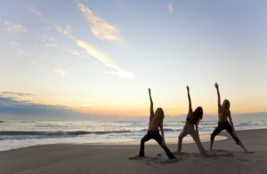 Three woman do yoga on the beach.